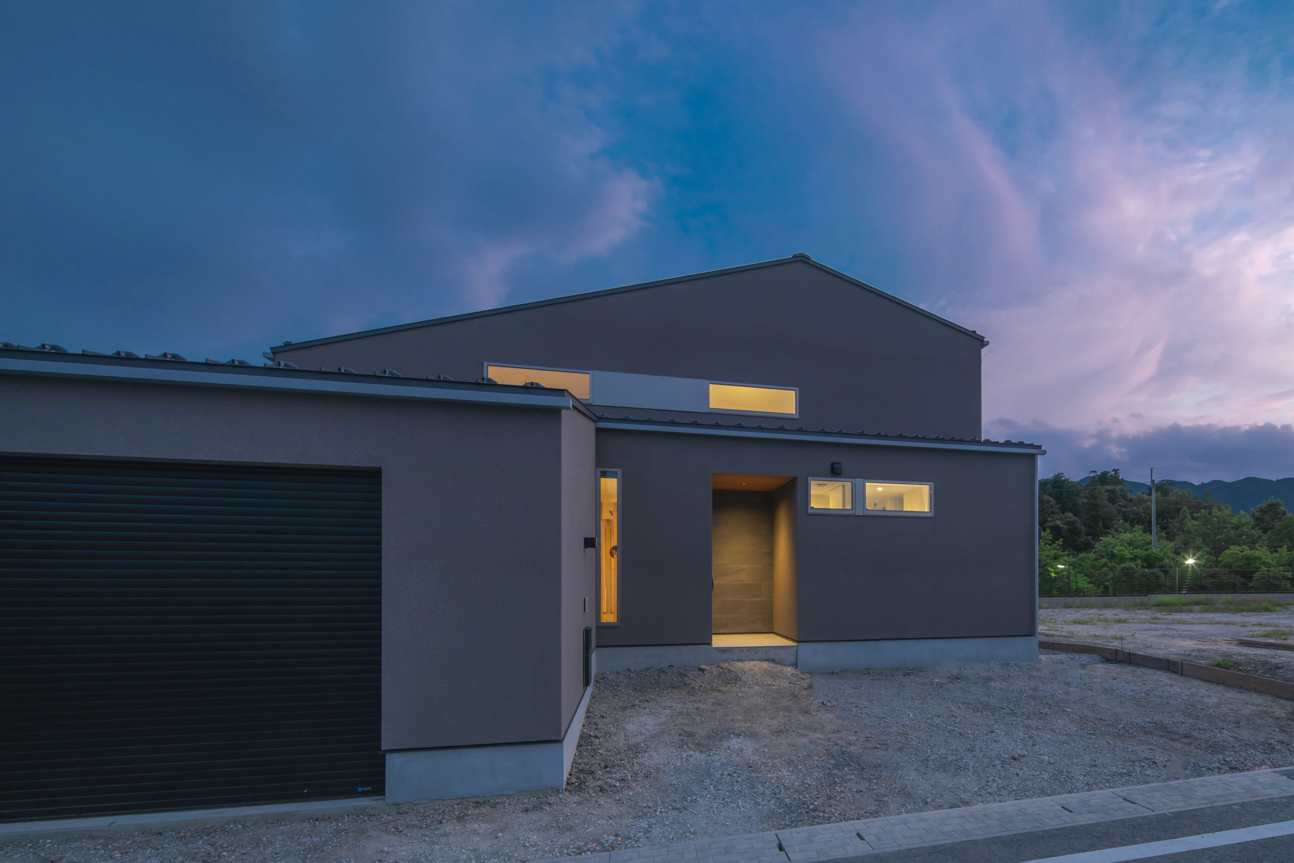 3Y1A7257 - 伊串工務店株式会社 Modern two-story gray house at dusk with illuminated entryway and windows, unfinished yard in foreground and hills in distance