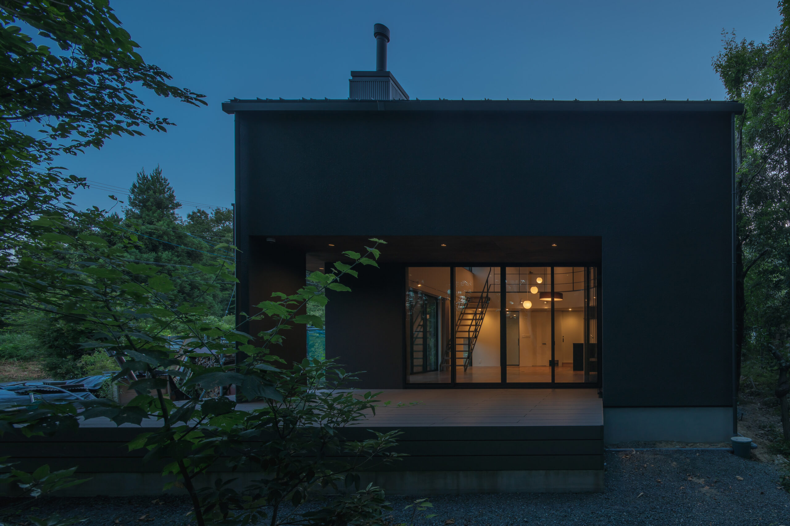 3Y1A7482 - 伊串工務店株式会社 Modern dark minimalist house with a large glass front, revealing a lit staircase inside, surrounded by trees at dusk.