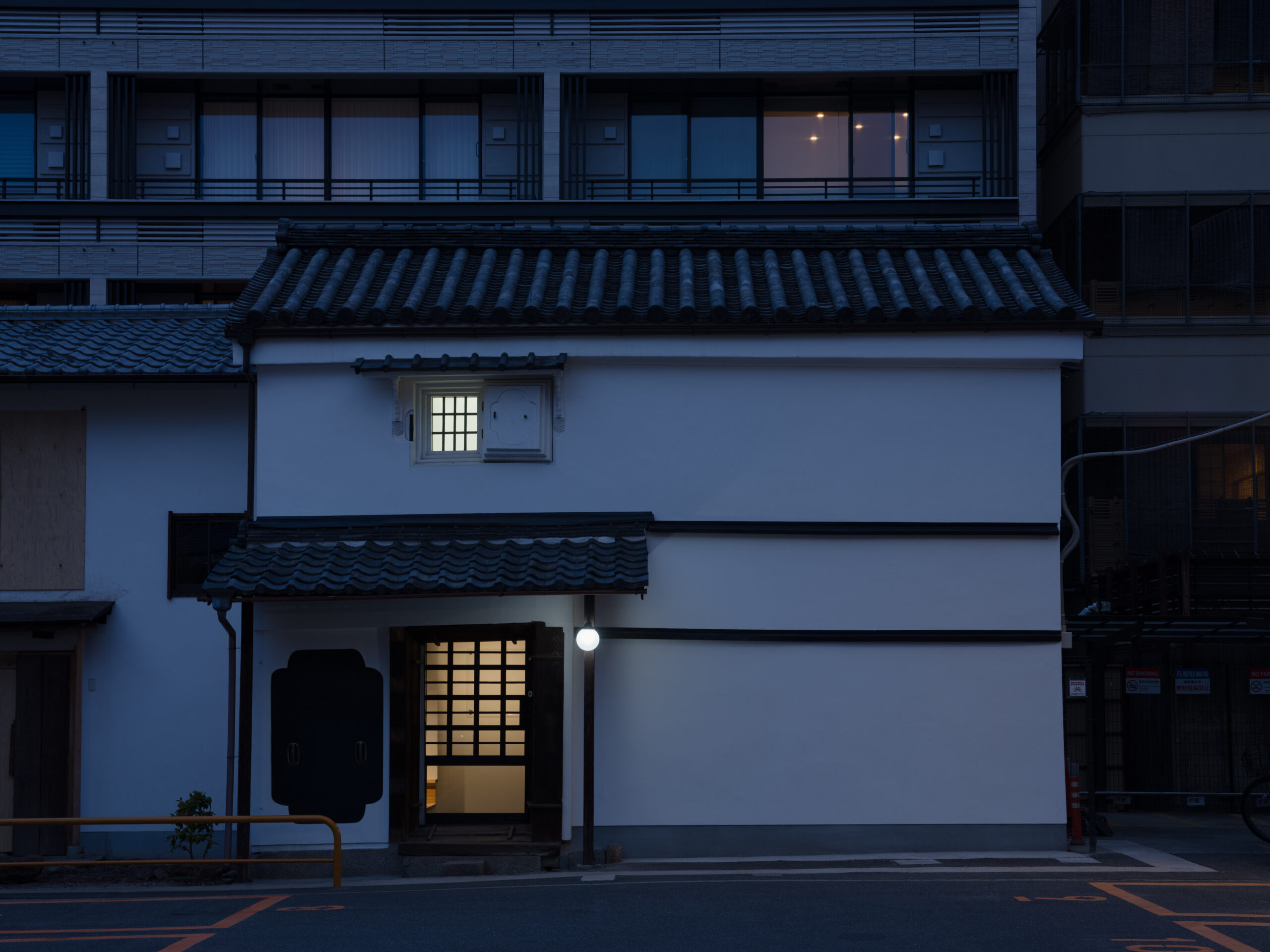 _DSF1900 - 伊串工務店株式会社 White building at dusk with a glowing doorway beneath a traditional tiled roof, blue evening light surrounding the façade.