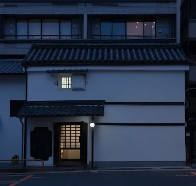 Traditional Japanese-style white storefront/house with dark tiled roof, lit doorway, at dusk.