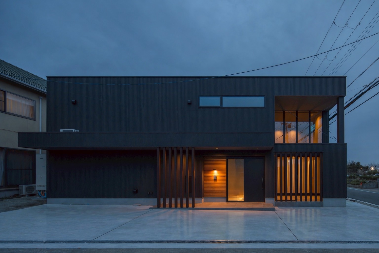 Modern dark gray two-story house at dusk with warm interior lights visible through large windows and wooden slatted entrance area
